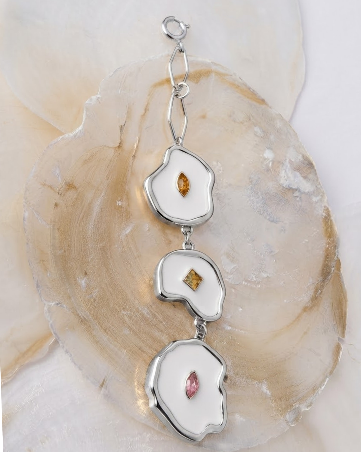 Silber Bag Charm -  Silberner Taschenanhänger, bestehend aus drei vertikal angeordneten, unregelmäßig geformten Elementen mit weißem Emaille, besetzt mit kleinen orangen, gelben und rosa Schmucksteinen, drapiert auf Perlmuttscheiben. 