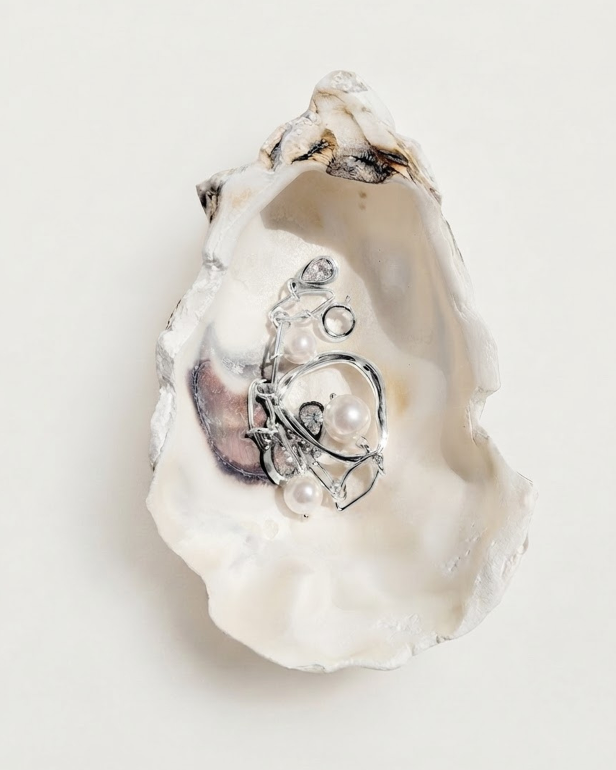 Silber Bag Charm -  Detailansicht einer geöffneten Austernschale, die als Schmuckschale dient und einen silbernen Anhänger verziert mit glänzenden Perlen und kleinen tropfenförmigen Schmucksteinen enthält.