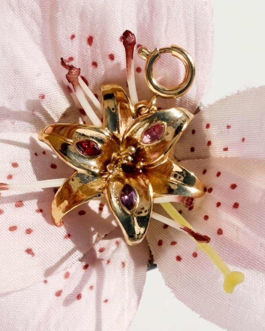 Gold BagBit -  Detailaufnahme eines goldenen Anhängers in Form einer Lilienblüte, besetzt mit drei kleinen Schmucksteinen in Rot, Rosa und Violett, drapiert auf einer rosafarbenen Lilie.