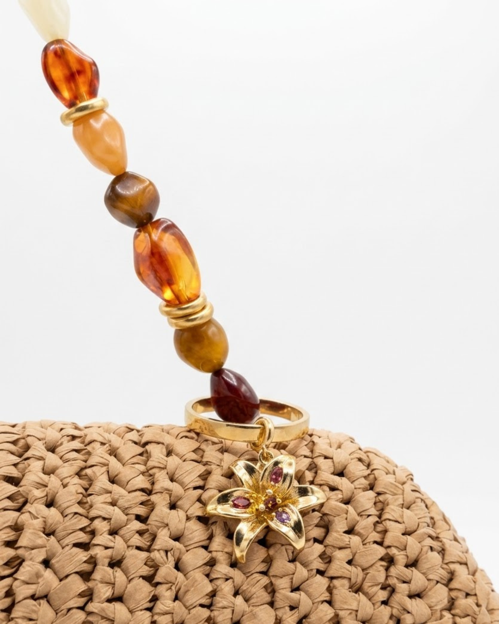 Gold BagBit -  Nahaufnahme einer geflochtenen Strohtasche, geschmückt mit einem Anhänger in Form einer goldenen Lilie, besetzt mit kleinen rosa, violetten und roten Schmucksteinen.