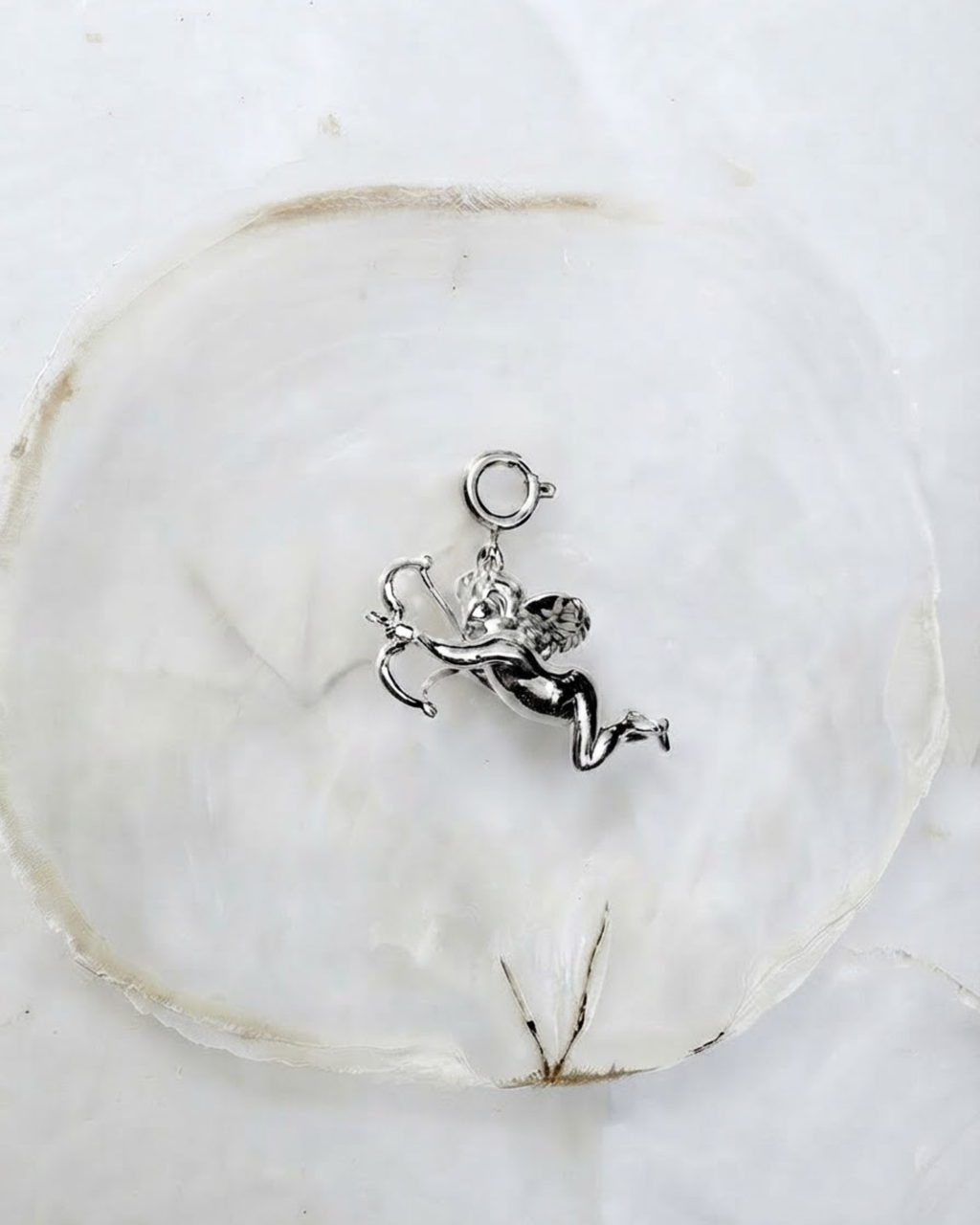 Silber Bag Bit - Silberfarbener Engel-Anhänger mit Pfeil und Bogen auf einer hellen Perlmutt-Unterlage.