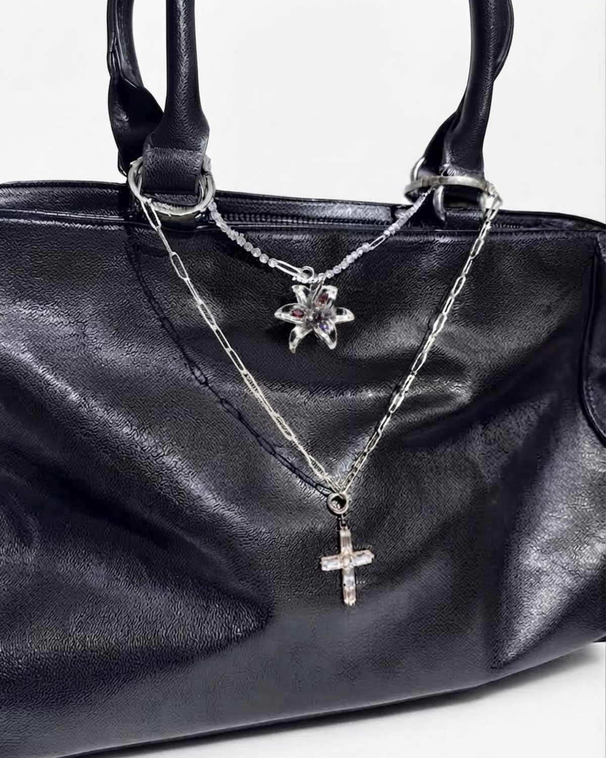 Silber Mixed Duo Chain - Schwarze Lederhandtasche mit zwei Halsketten zwischen den Henkeln: eine obere silberne Strasskette mit einer silbernen Blume-Anhänger angehangen und eine untere silberne Gliederkette mit einem Kristallkreuz-Anhänger.