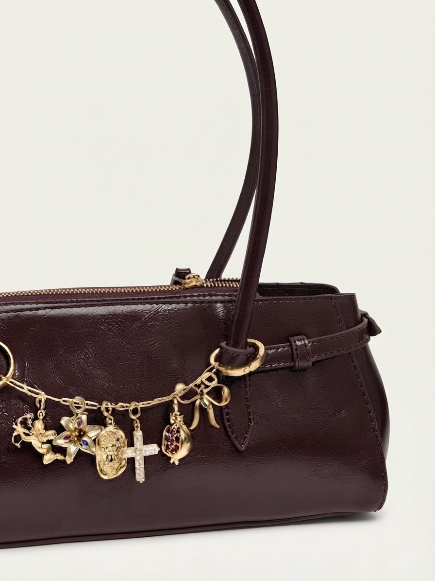 Gold Bag Chain - Dunkelrote Schultertasche aus glänzendem Leder, verziert mit einer goldfarbenen Kette zwischen den Henkeln und an dieser befinden sich mehrere goldene Anhängern (Motiv: Engel, Blumen, Kreuz, Schleife, Teddybär-Relief und Granatapfel).