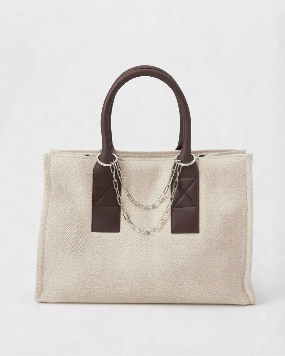 Silber Bag Chain - Beige-cremefarbene Tasche, verziert mit braunen Ledergriffen und einer silberfarbene Kette, die zwischen den Henkeln gespannt ist.