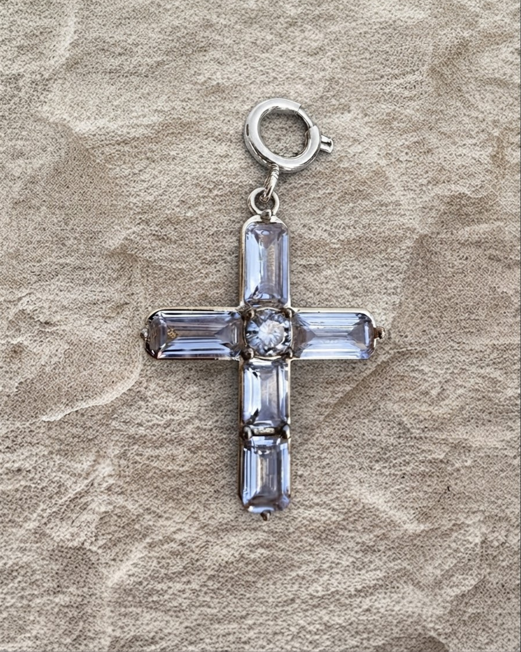 Silber BagBit -  Goldener Kreuz-Anhänger, besetzt mit vier länglichen und einem zentralen runden, klaren Schmuckstein, liegend auf einer sandsteinfarbenen Oberfläche.