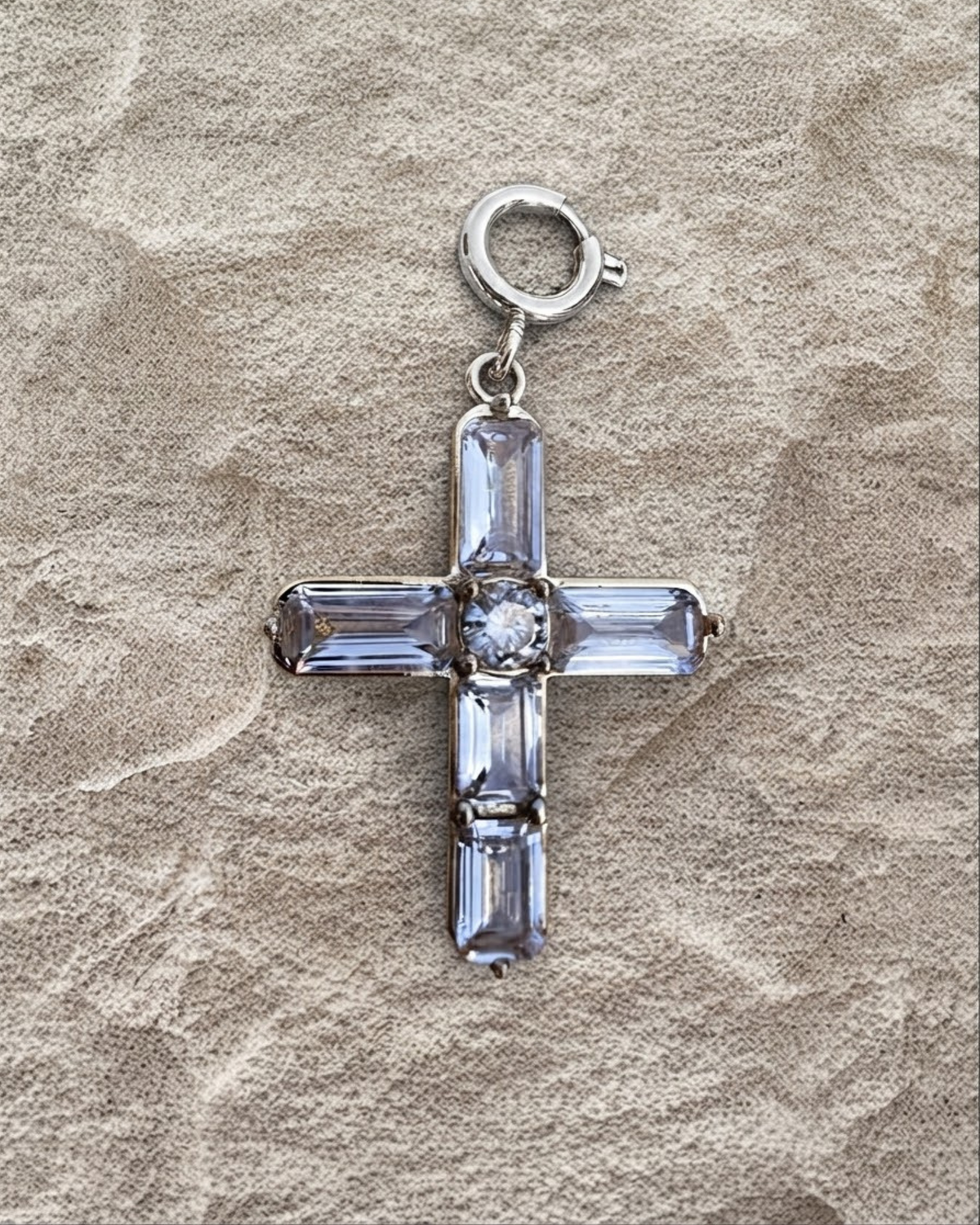 Silber BagBit -  Goldener Kreuz-Anhänger, besetzt mit vier länglichen und einem zentralen runden, klaren Schmuckstein, liegend auf einer sandsteinfarbenen Oberfläche.