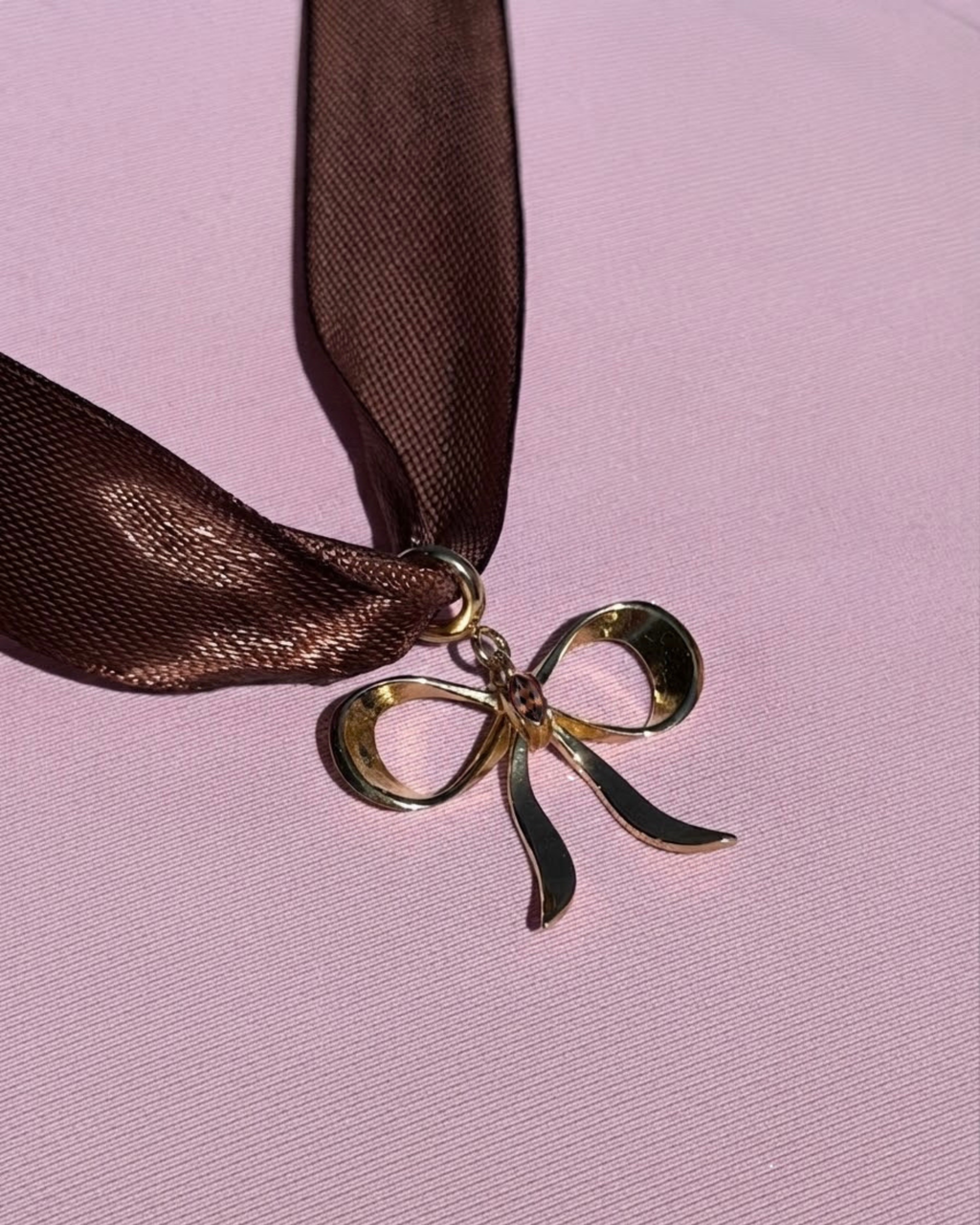 Gold BagBit - Nahaufnahme eines braunen Seidenbands und einem Goldanhänger in Form einer Schleife, drapiert auf einem strukturierten rosa Hintergrund.