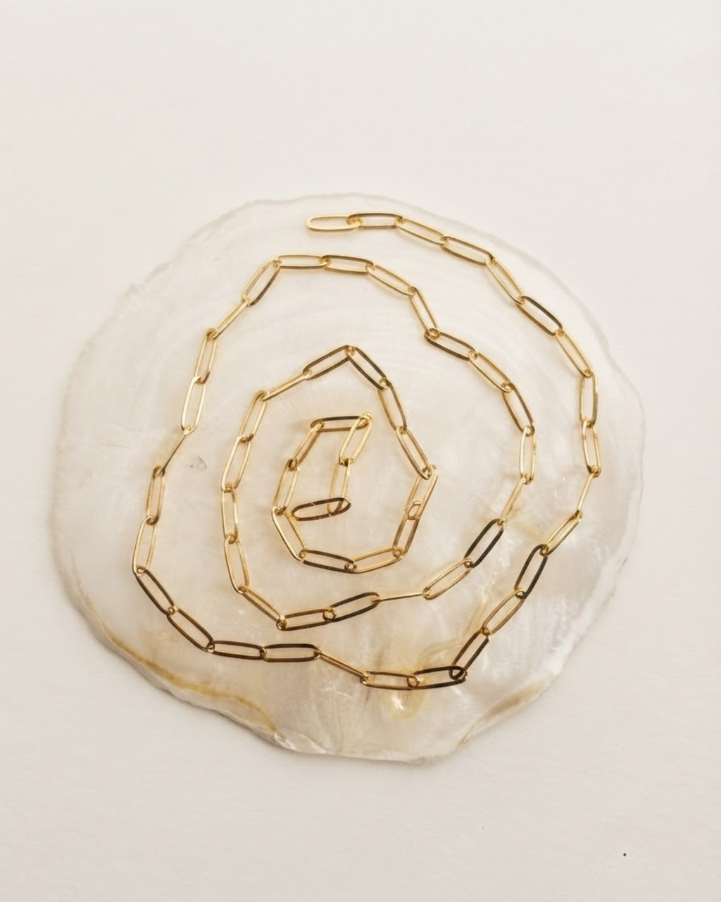 Gold Single Chain - Eine spiralförmig auf einer hellen Perlmutt-Scheibe arrangierte, goldfarbene Kette mit länglichen Gliedern.