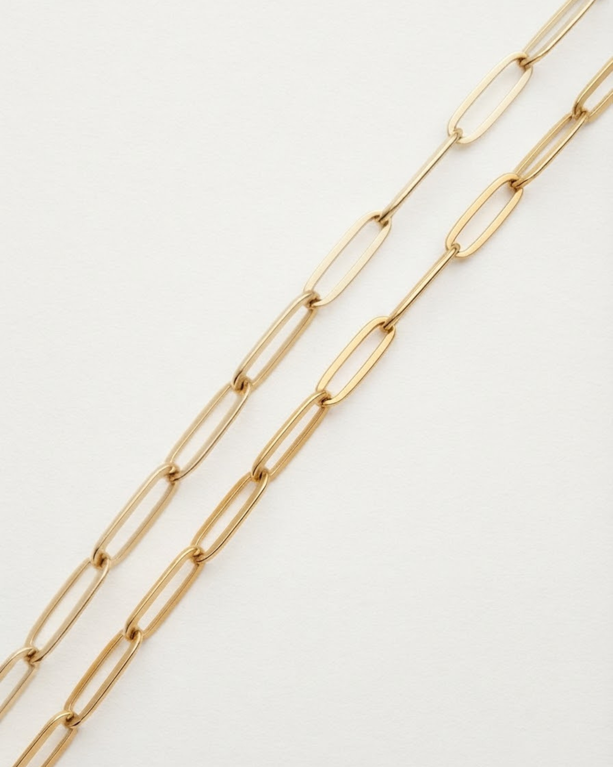 Gold Bag Chain - Nahaufnahme von zwei parallelen, goldfarbenen Ketten mit länglichen Gliedern, die diagonal über einen weißen Hintergrund verlaufen.