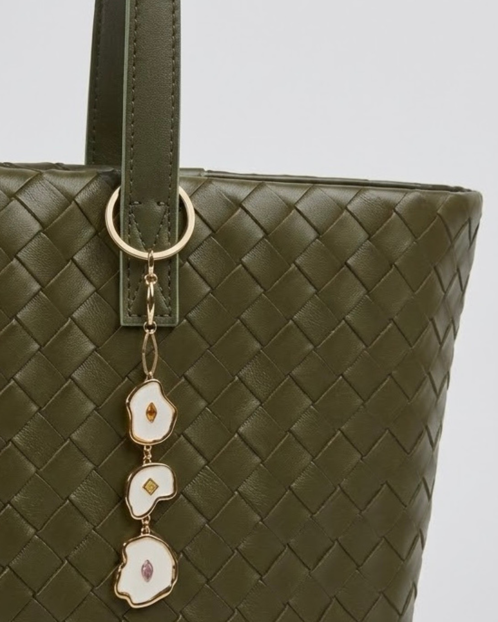 Gold Bag Charm-  Nahaufnahme einer olivgrünen, geflochtenen Leder-Handtasche, verziert mit einem herabhängenden Anhänger aus drei unregelmäßig geformten, goldumrandeten Emaille-Elementen mit kleinen Schmucksteinen.