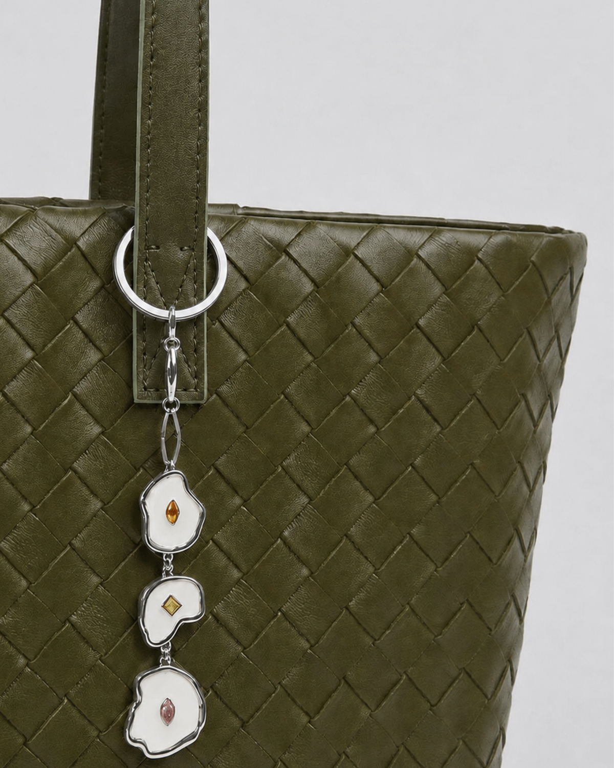Silber Bag Charm -  Nahaufnahme einer olivgrünen, geflochtenen Leder-Handtasche, verziert mit einem herabhängenden Anhänger aus drei unregelmäßig geformten, silberumrandeten Emaille-Elementen mit kleinen Schmucksteinen.