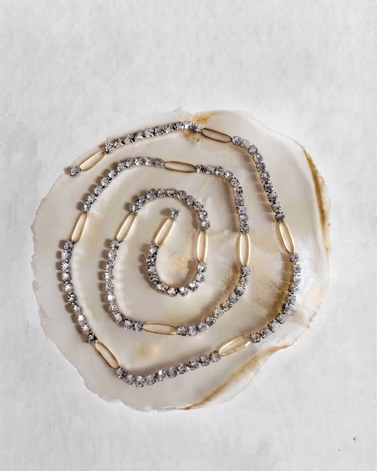 Gold Strass Single Chain - Eine spiralförmig auf einer hellen Perlmutt-Scheibe arrangierte Strasskette mit klaren Kristallen, die in regelmäßigen Abständen durch glatte, goldfarbene ovale Glieder unterbrochen wird.