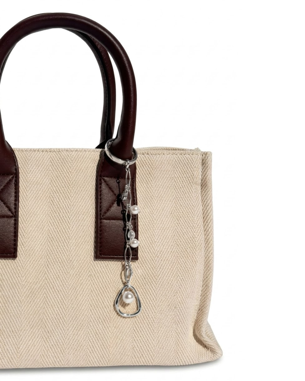Silber Bag Charm -  Aufnahme einer beigefarbenen Canvas-Tragetasche mit Griffen aus dunkelbraunem Leder und einem filigranen silbernen Anhänger, verziert mit Perlen.