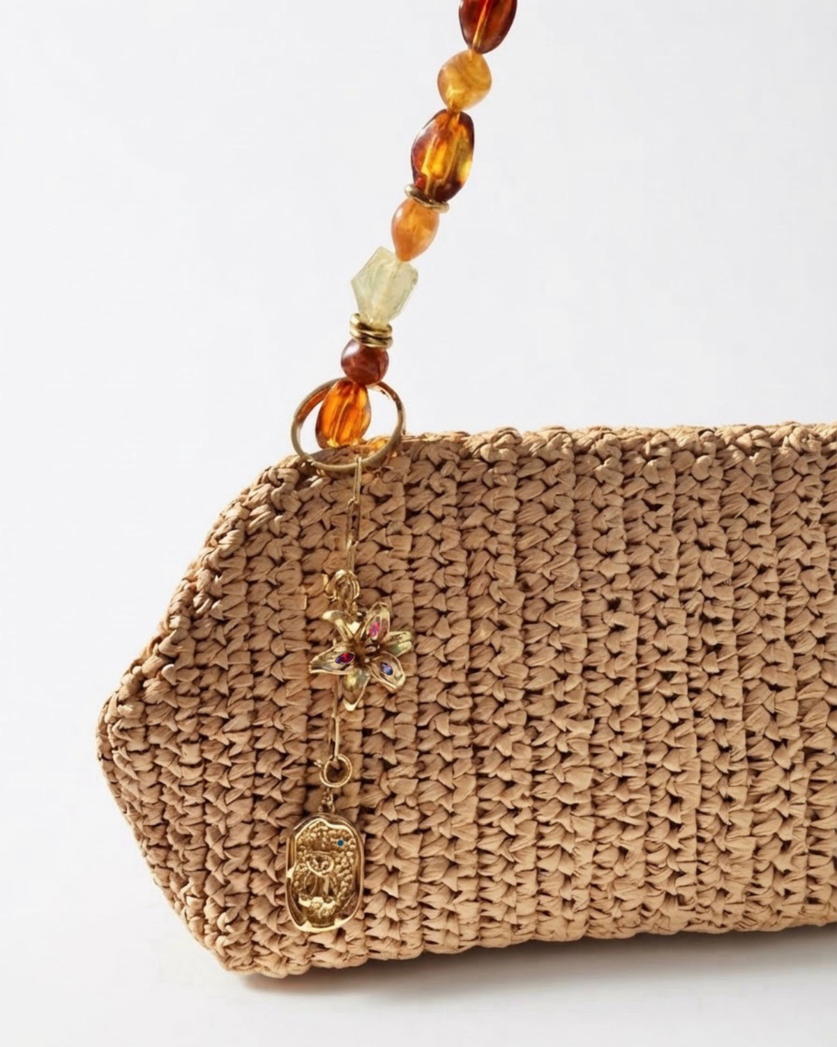 Gold Bad Bit - Beigefarbene Tasche mit einer kurzen goldenen Kette mit Blumen- und Medaillon-Anhänger am Taschenhenkel befestigt.