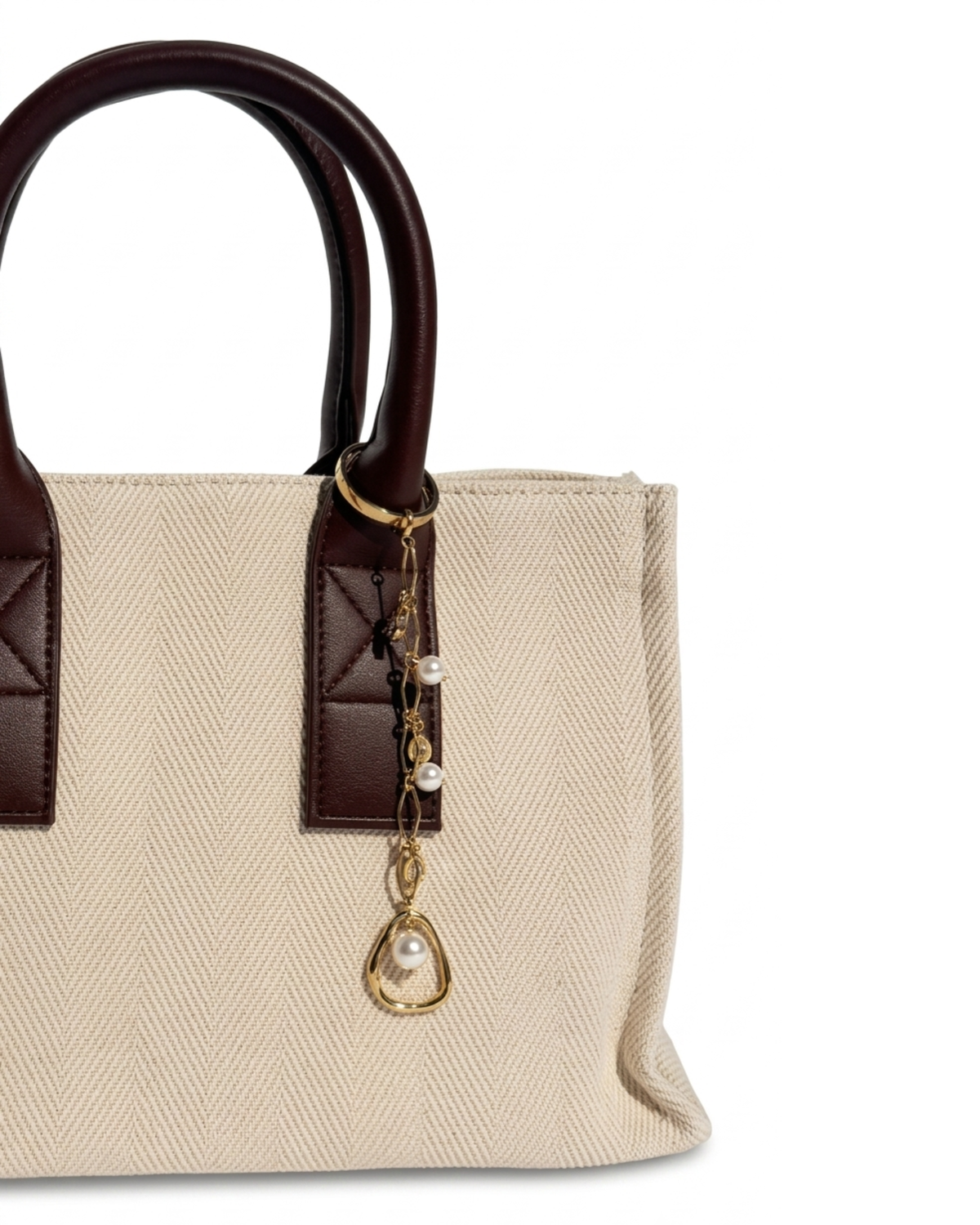 Gold Bag Charm -  Aufnahme einer beigefarbenen Canvas-Tragetasche mit Griffen aus dunkelbraunem Leder und einem filigranen goldenen Anhänger, verziert mit Perlen.