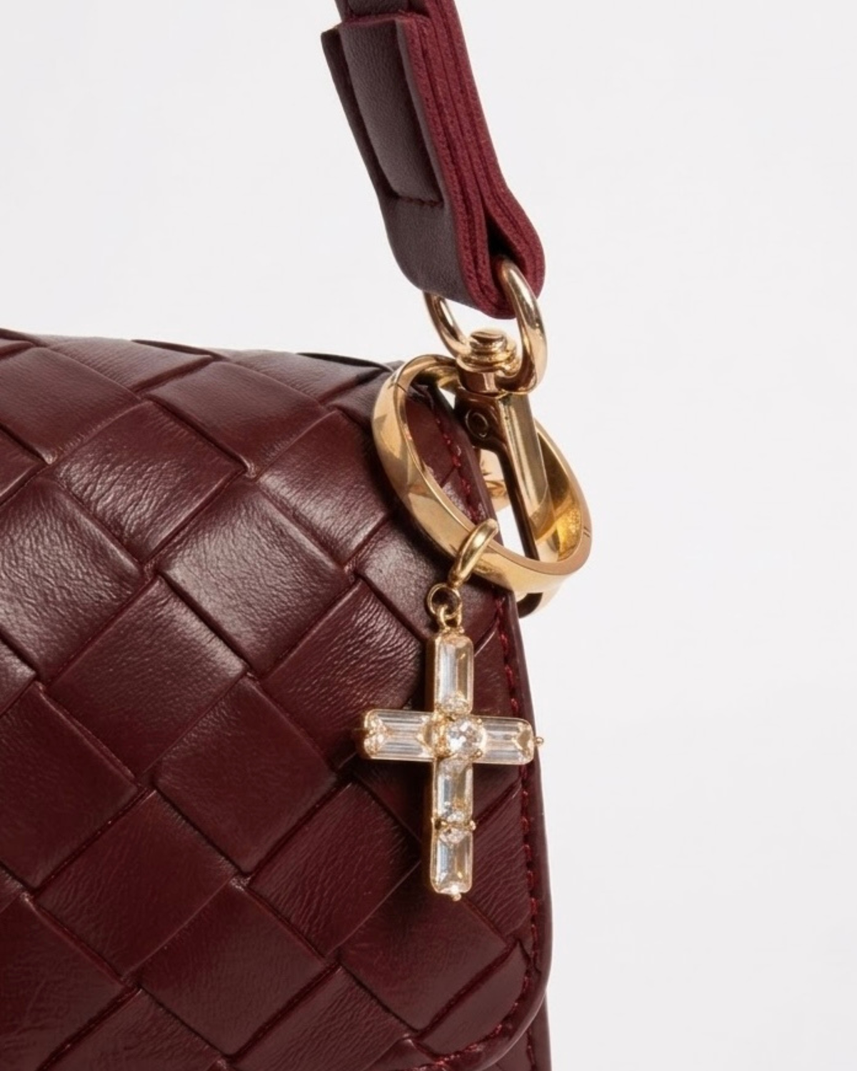 Gold BagBit -  Nahaufnahme einer weinroten, geflochtenen Ledertasche, an der ein Ring mit einem goldenen Anhänger in Form eines Kreuzes hängt, der mit klaren Schmucksteinen besetzt ist.