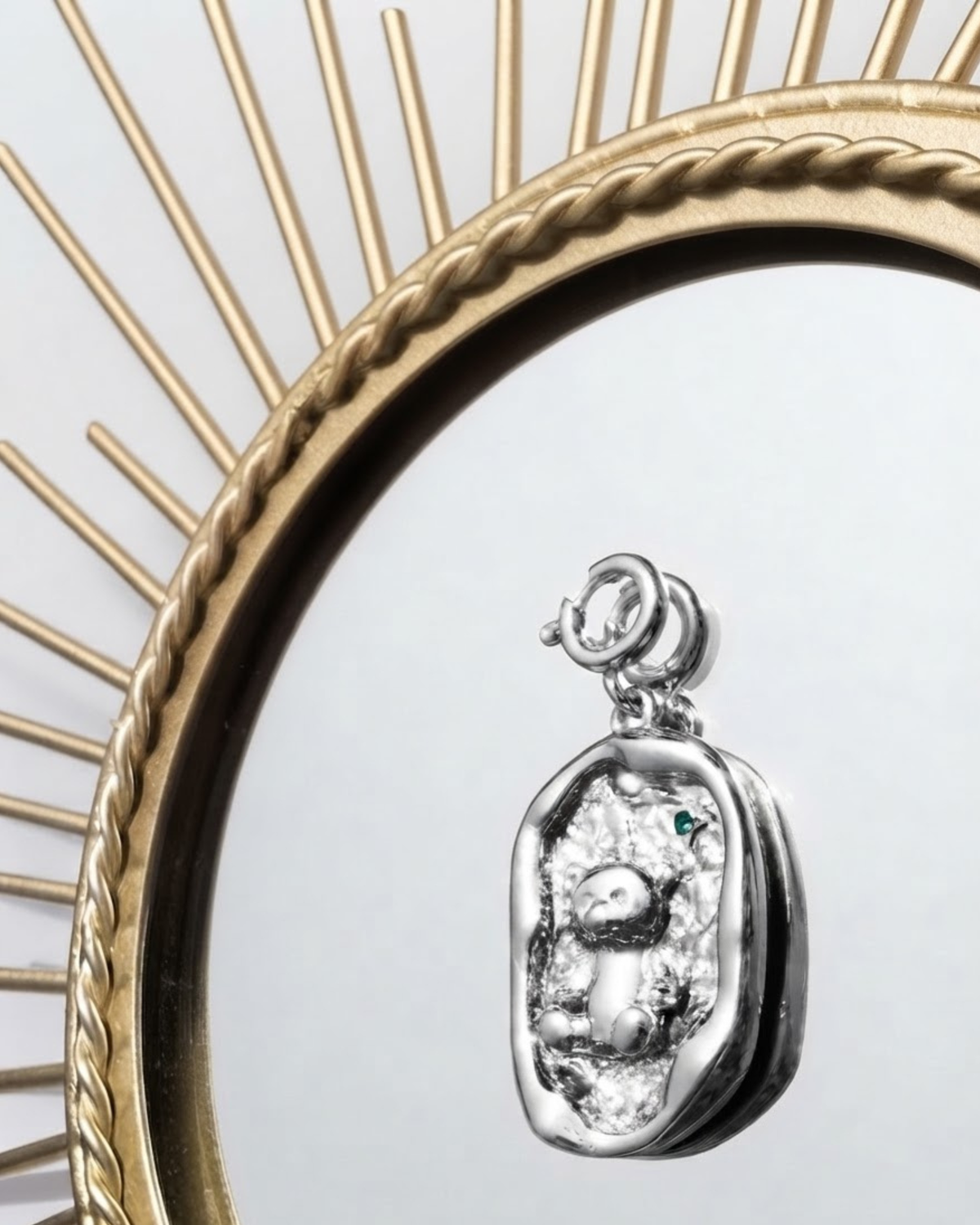 Silber BagBit - Detailaufnahme eines silbernen Anhängers mit unregelmäßigem Rand, der einen Teddybären auf einer strukturierten Fläche zeigt und von einem goldenen, sonnenförmigen Spiegelrahmen umgeben ist.