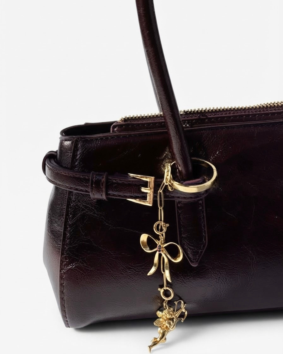 Gold Bag Bit - Dunkelrote Schultertasche aus glänzendem Leder mit einer kurzen, herabhängenden goldenen Kette, dekoriert mit einer Schleife und einem Engel-Anhänger.