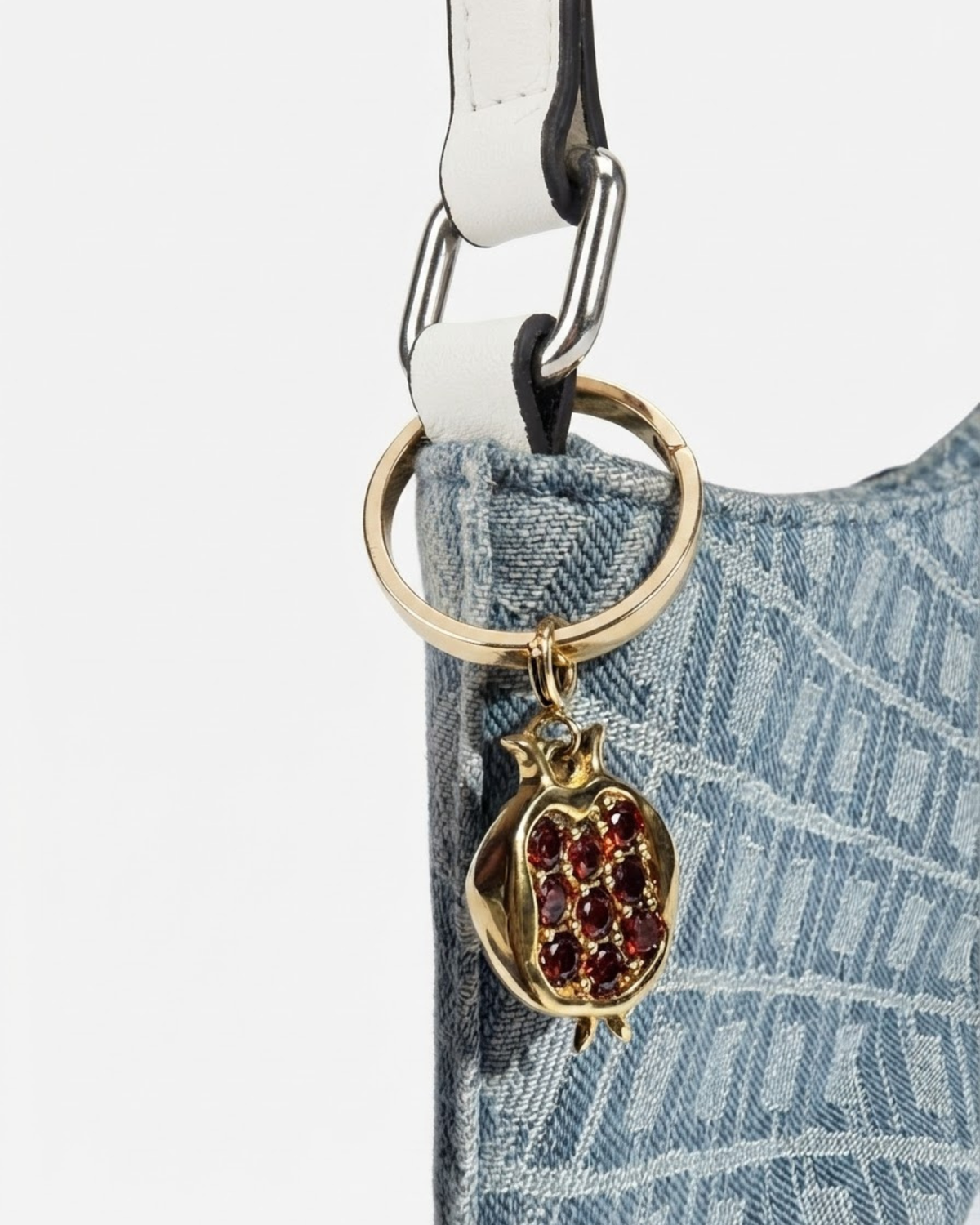 Gold Bag Bit - Nahaufnahme einer blau-gemusterte Denim-Tasche mit einem weißen Schulterriemen und daran einen goldenen Granatapfel-Anhänger befestigt.