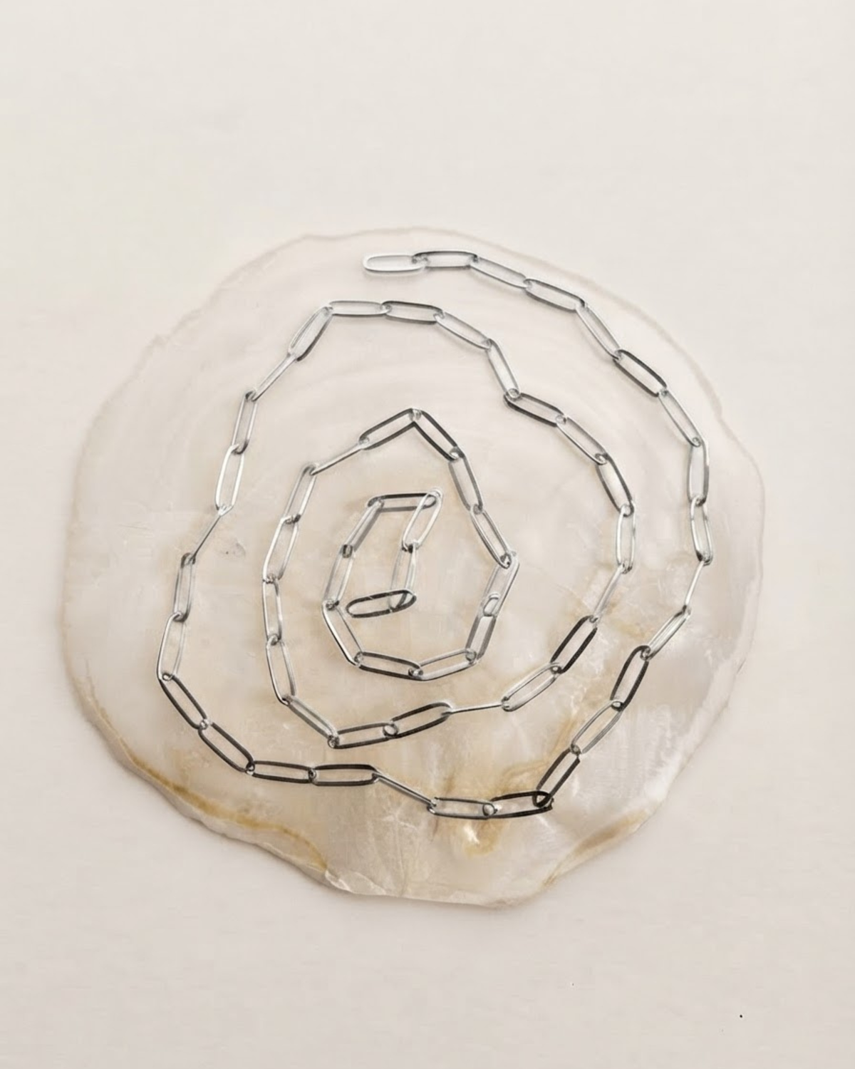 Silber Bag Chain - Eine spiralförmig auf einer hellen Perlmutt-Scheibe arrangierte, silberfarbenen Kette mit länglichen Gliedern.