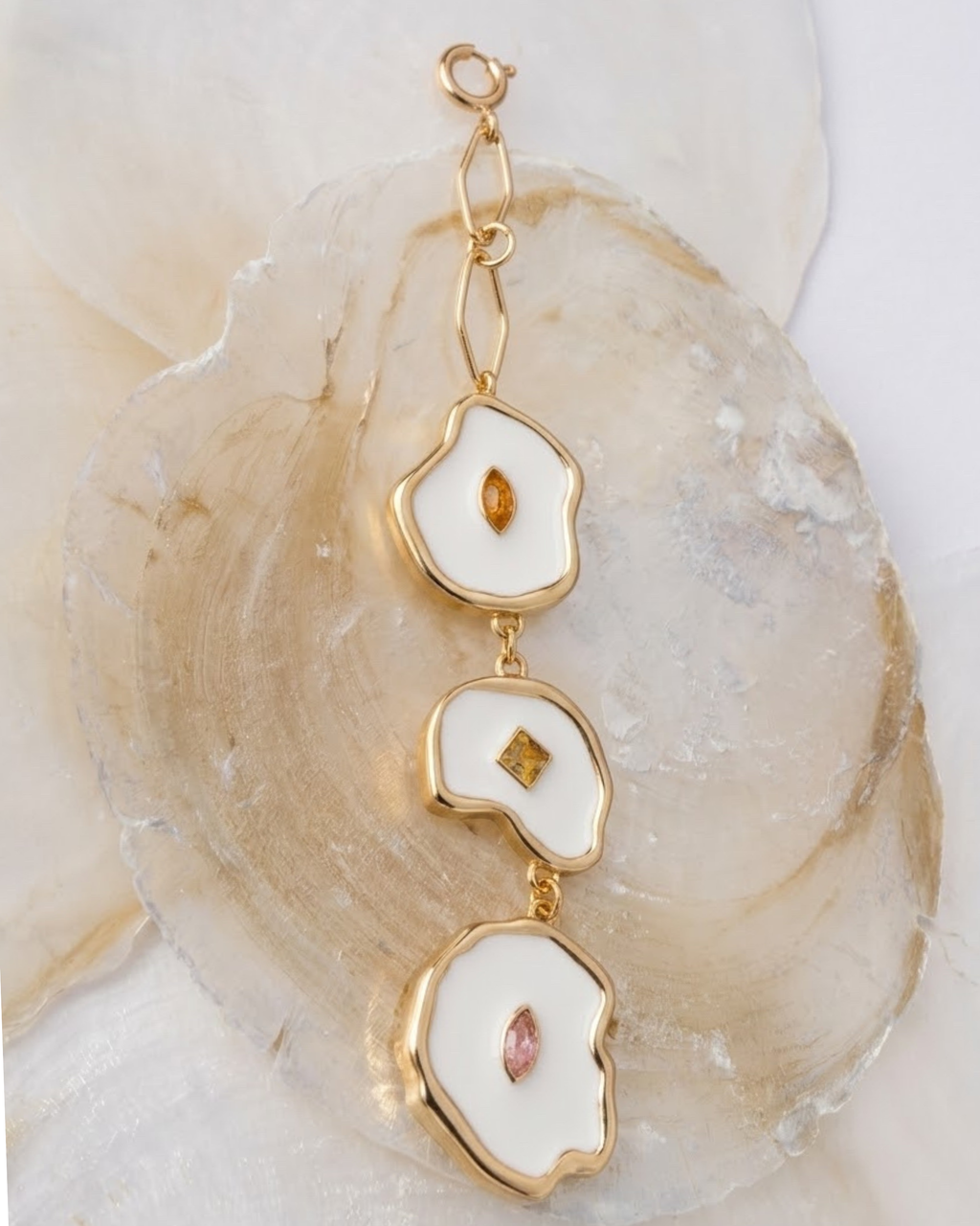 Gold Bag Charm -  Goldener Taschenanhänger, bestehend aus drei vertikal angeordneten, unregelmäßig geformten Elementen mit weißem Emaille, besetzt mit kleinen orangen, gelben und rosa Schmucksteinen, drapiert auf Perlmuttscheiben.