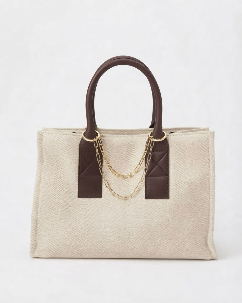 Gold Bag Chain - Beige-cremefarbene Tasche, verziert mit braunen Ledergriffen und einer goldfarbenen Kette, die zwischen den Henkeln gespannt ist.