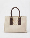 Gold Bag Chain - Beige-cremefarbene Tasche, verziert mit braunen Ledergriffen und einer goldfarbenen Kette, die zwischen den Henkeln gespannt ist.