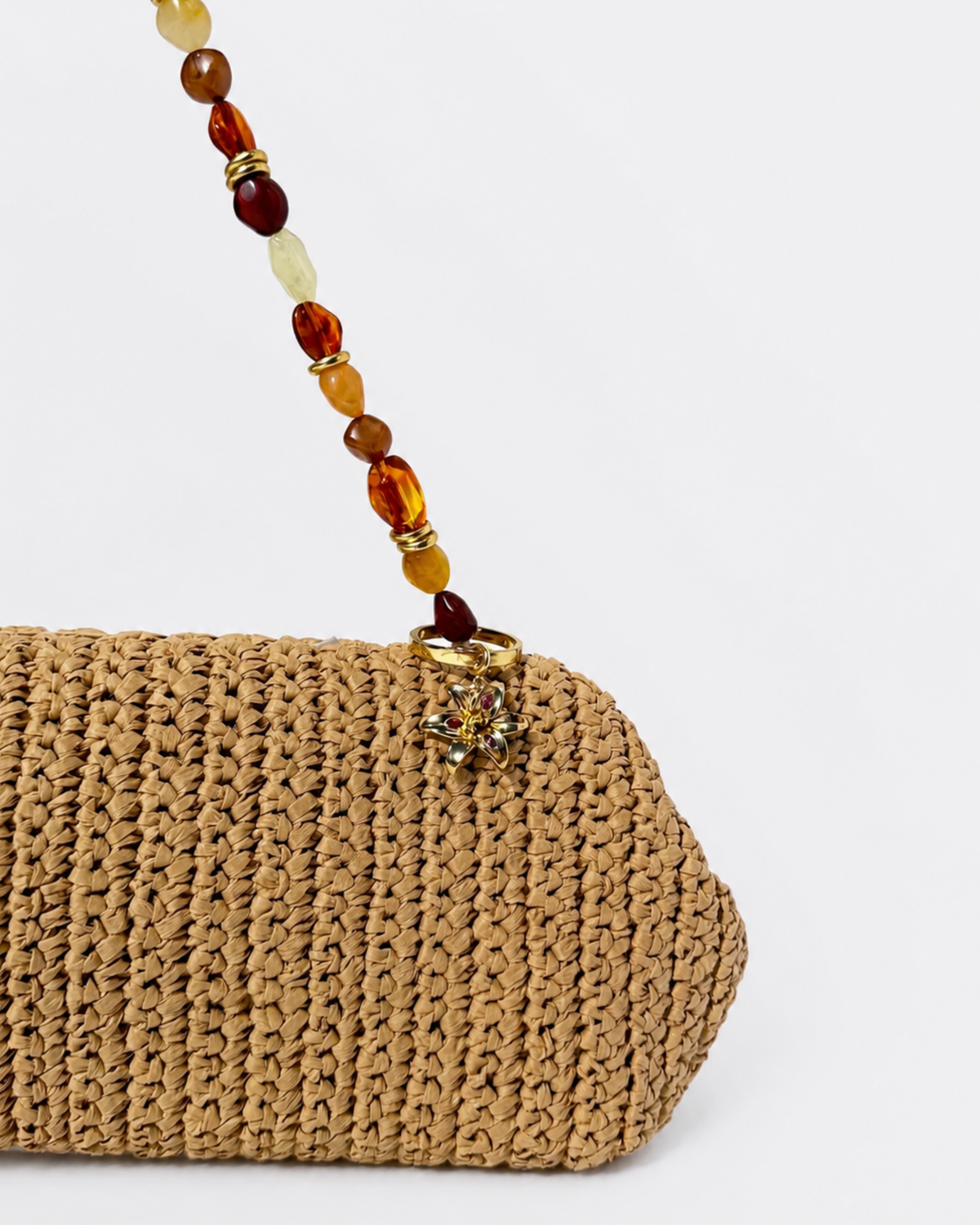 Gold BagBit -  Aufnahme einer geflochtenen Strohtasche, geschmückt mit einem Anhänger in Form einer goldenen Lilie, besetzt mit kleinen rosa, violetten und roten Schmucksteinen.