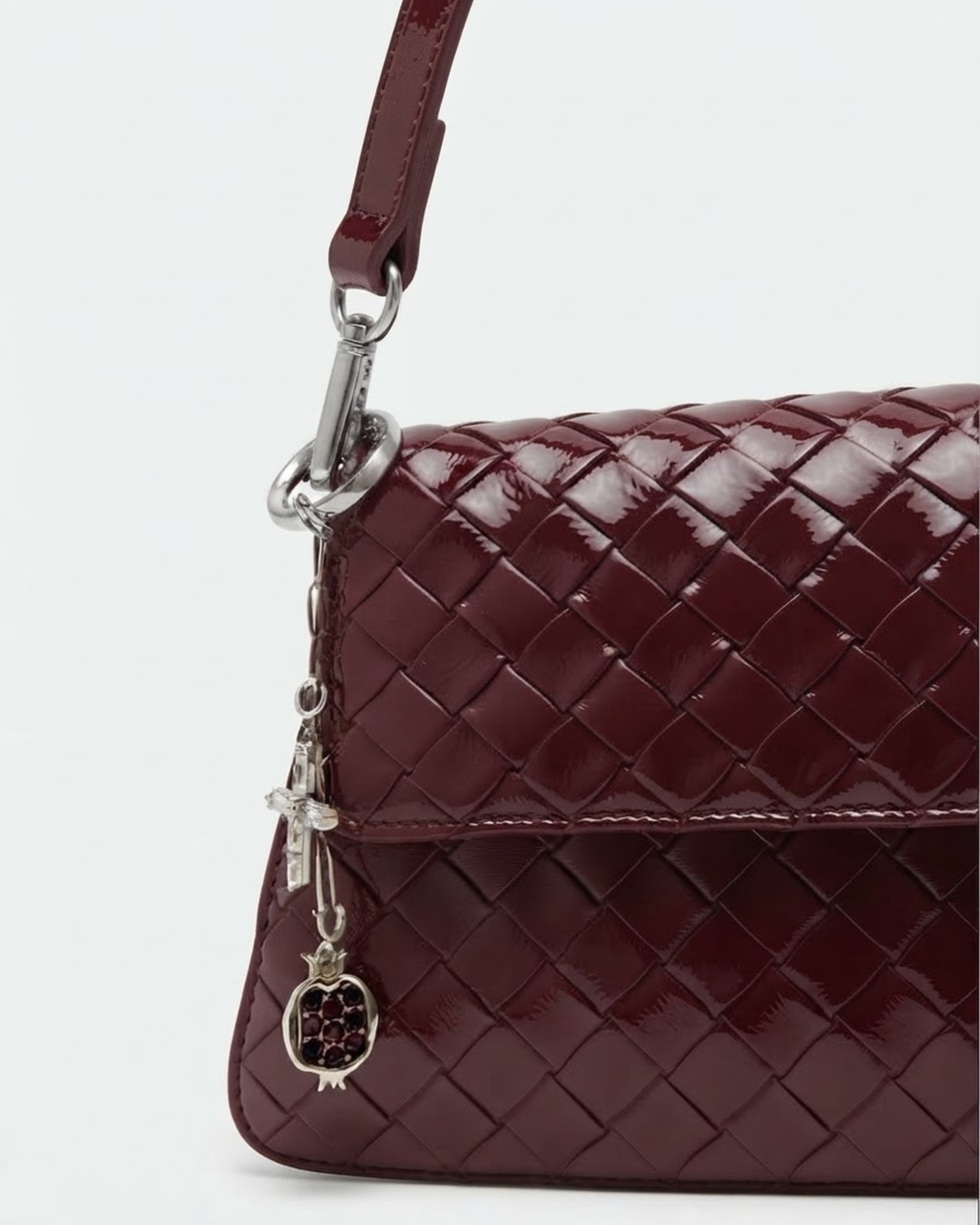 Silber Bad Bit - Dunkelrot geflochtene Ledertasche mit glänzender Oberfläche und einer kurzen silbernen Kette herabhängend am Taschenhenkel, verziert mit einem Kreuz- und einem Granatapfel-Anhänger.