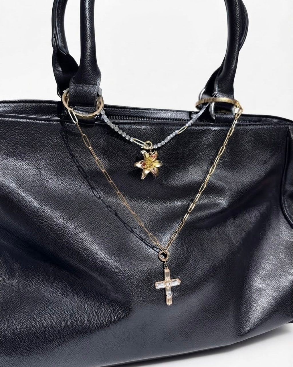Gold Mixed Duo Chain - Schwarze Lederhandtasche mit zwei Halsketten zwischen den Henkeln: eine obere goldene Strasskette mit einer goldenen Blume-Anhänger angehangen und eine untere goldene Gliederkette mit einem Kristallkreuz-Anhänger.