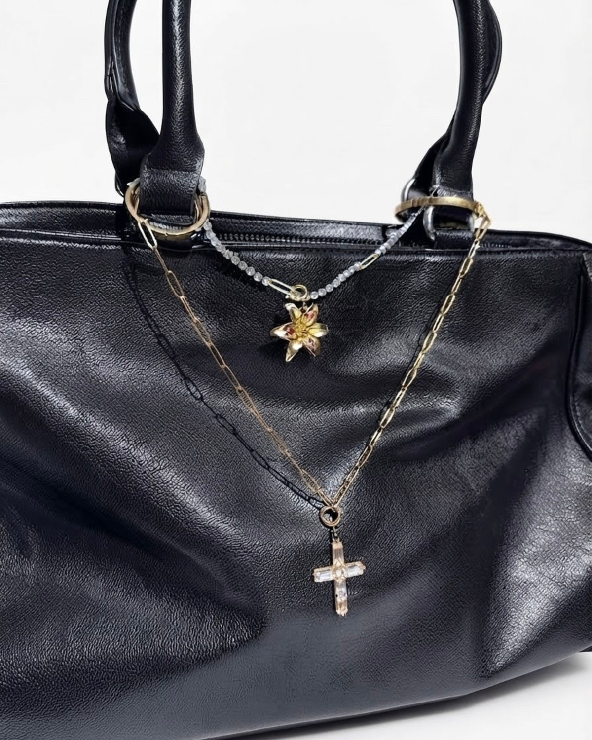 Gold Mixed Duo Chain - Schwarze Lederhandtasche mit zwei Halsketten zwischen den Henkeln: eine obere goldene Strasskette mit einer goldenen Blume-Anhänger angehangen und eine untere goldene Gliederkette mit einem Kristallkreuz-Anhänger.