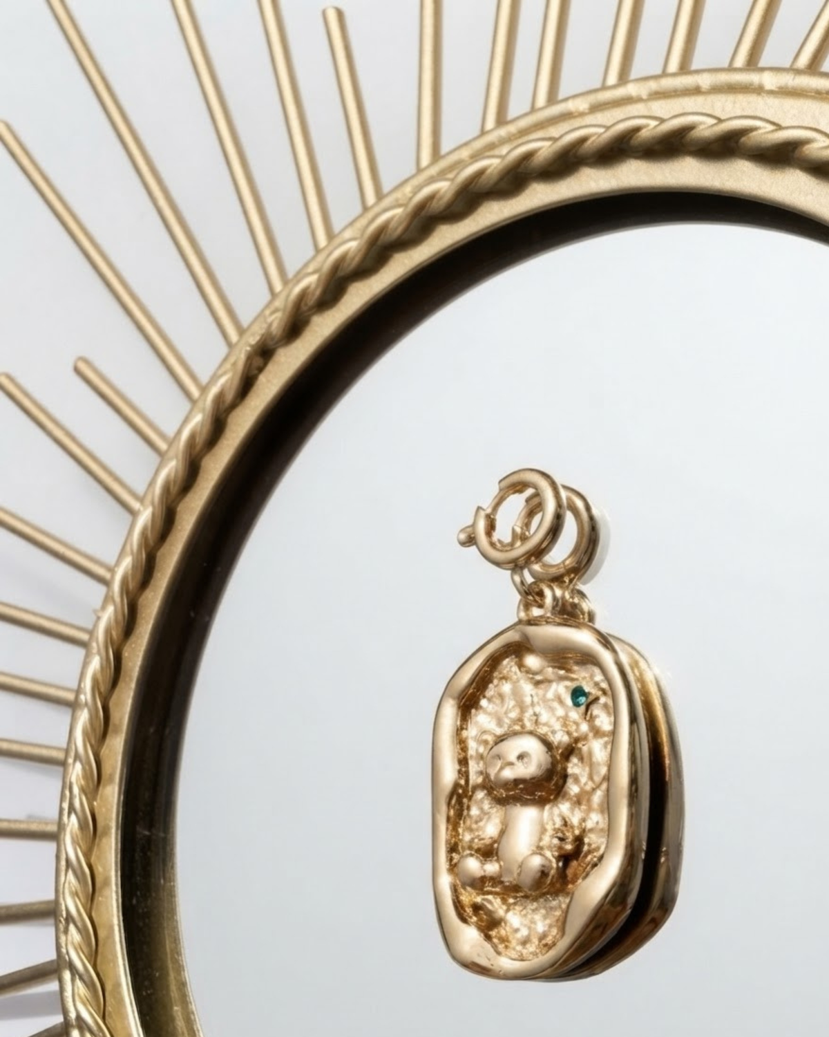 Gold BagBit - Detailaufnahme eines goldenen Anhängers mit unregelmäßigem Rand, der einen Teddybären auf einer strukturierten Fläche zeigt und von einem goldenen, sonnenförmigen Spiegelrahmen umgeben ist.