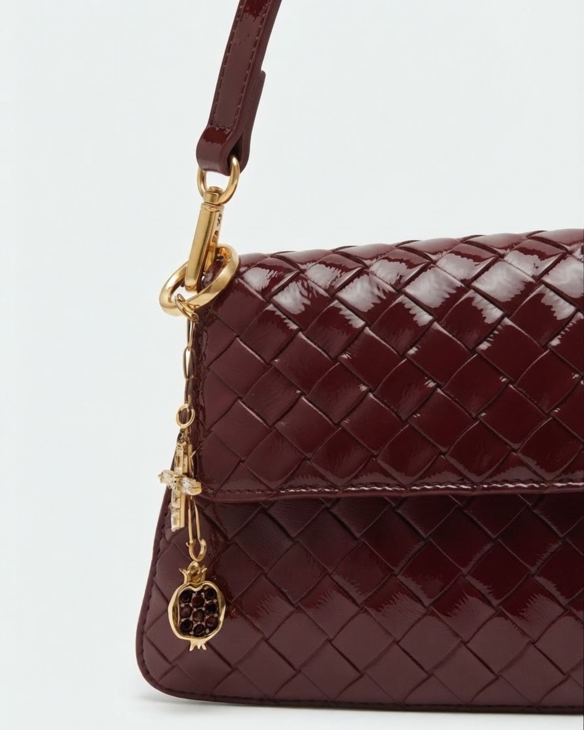 Gold Bag Bit - Dunkelrot geflochtene Ledertasche mit glänzender Oberfläche und einer kurzen goldenen Kette herabhängend am Taschenhenkel, verziert mit einem Kreuz- und einem Granatapfel-Anhänger.