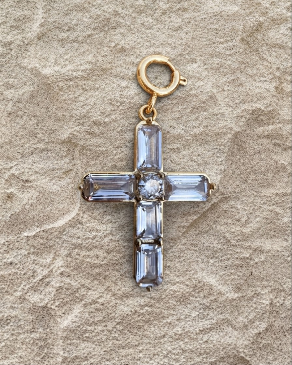 Gold BagBit -  Goldener Kreuz-Anhänger, besetzt mit vier länglichen und einem zentralen runden, klaren Schmuckstein, liegend auf einer sandsteinfarbenen Oberfläche.