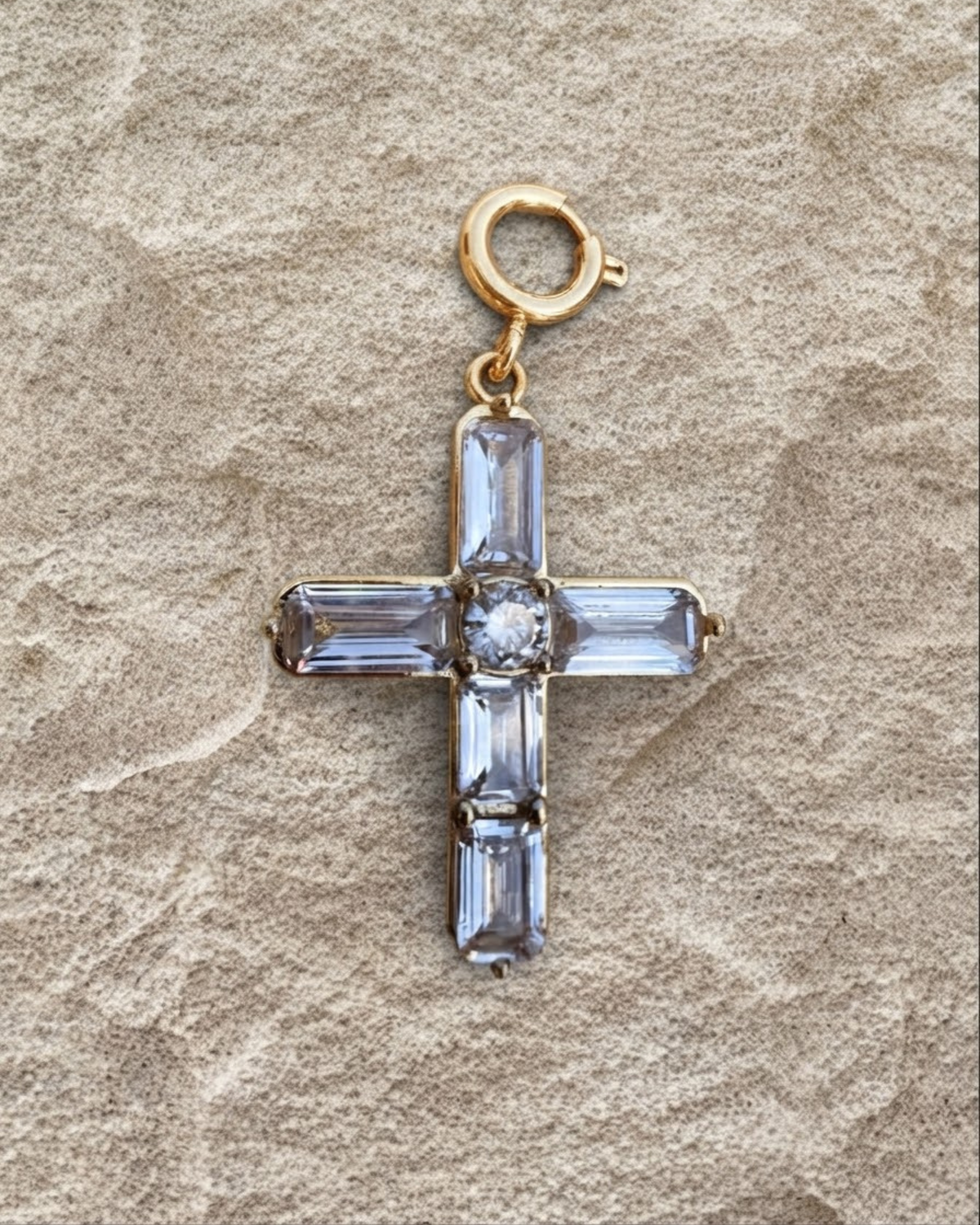 Gold BagBit -  Goldener Kreuz-Anhänger, besetzt mit vier länglichen und einem zentralen runden, klaren Schmuckstein, liegend auf einer sandsteinfarbenen Oberfläche.