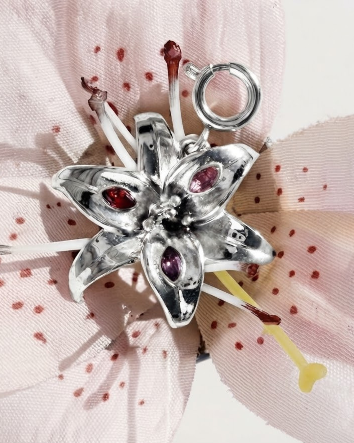 Silber BagBit -  Detailaufnahme eines silbernen Anhängers in Form einer Lilienblüte, besetzt mit drei kleinen Schmucksteinen in Rot, Rosa und Violett, drapiert auf einer rosafarbenen Lilie.