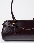 Gold Bag Chain - Dunkelrote Schultertasche aus glänzendem Leder mit einer feinen, goldfarbenen Gliederkette als Detail zwischen den beiden Henkeln gespannt.