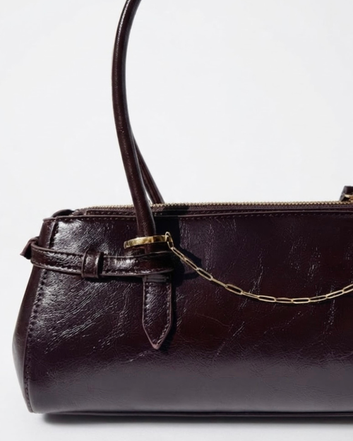 Gold Bag Chain - Dunkelrote Schultertasche aus glänzendem Leder mit einer feinen, goldfarbenen Gliederkette als Detail zwischen den beiden Henkeln gespannt.