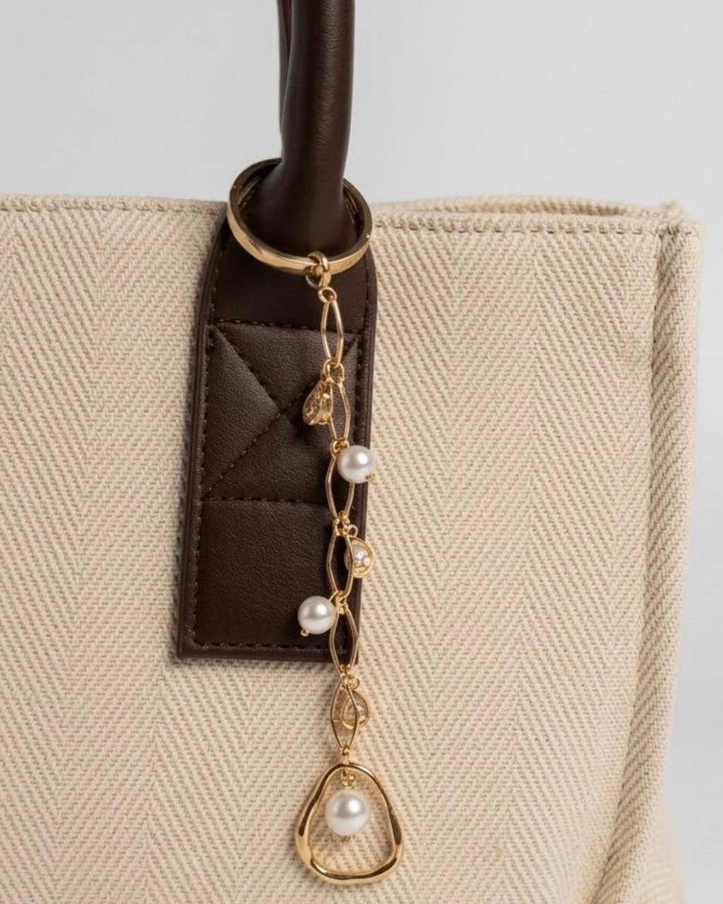 Gold Bag Charm -  Nahaufnahme einer beigefarbenen Canvas-Tragetasche mit braunem Ledergriff und einem filigranen goldenen Anhänger, der mit Perlen und tropfenförmigen Schmucksteinen verziert ist.