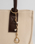 Gold Bag Charm -  Nahaufnahme einer beigefarbenen Canvas-Tragetasche mit braunem Ledergriff und einem filigranen goldenen Anhänger, der mit Perlen und tropfenförmigen Schmucksteinen verziert ist.