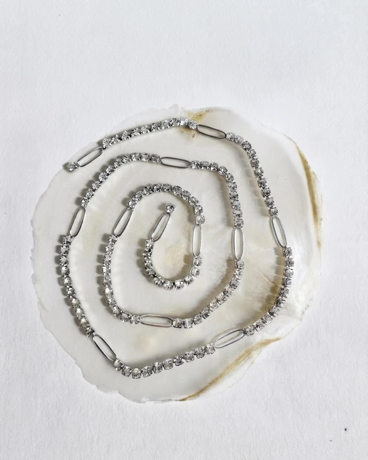 Silber Strass Duo Chain - Eine spiralförmig auf einer hellen Perlmutt-Scheibe arrangierte Kette mit klaren Kristallen, die in regelmäßigen Abständen durch glatte, silberfarbene ovale Glieder unterbrochen wird.