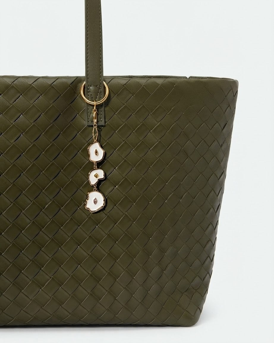 Gold Bag Charm -  Aufnahme einer olivgrünen, geflochtenen Leder-Handtasche, verziert mit einem herabhängenden Anhänger aus drei unregelmäßig geformten, goldumrandeten Emaille-Elementen mit kleinen Schmucksteinen.