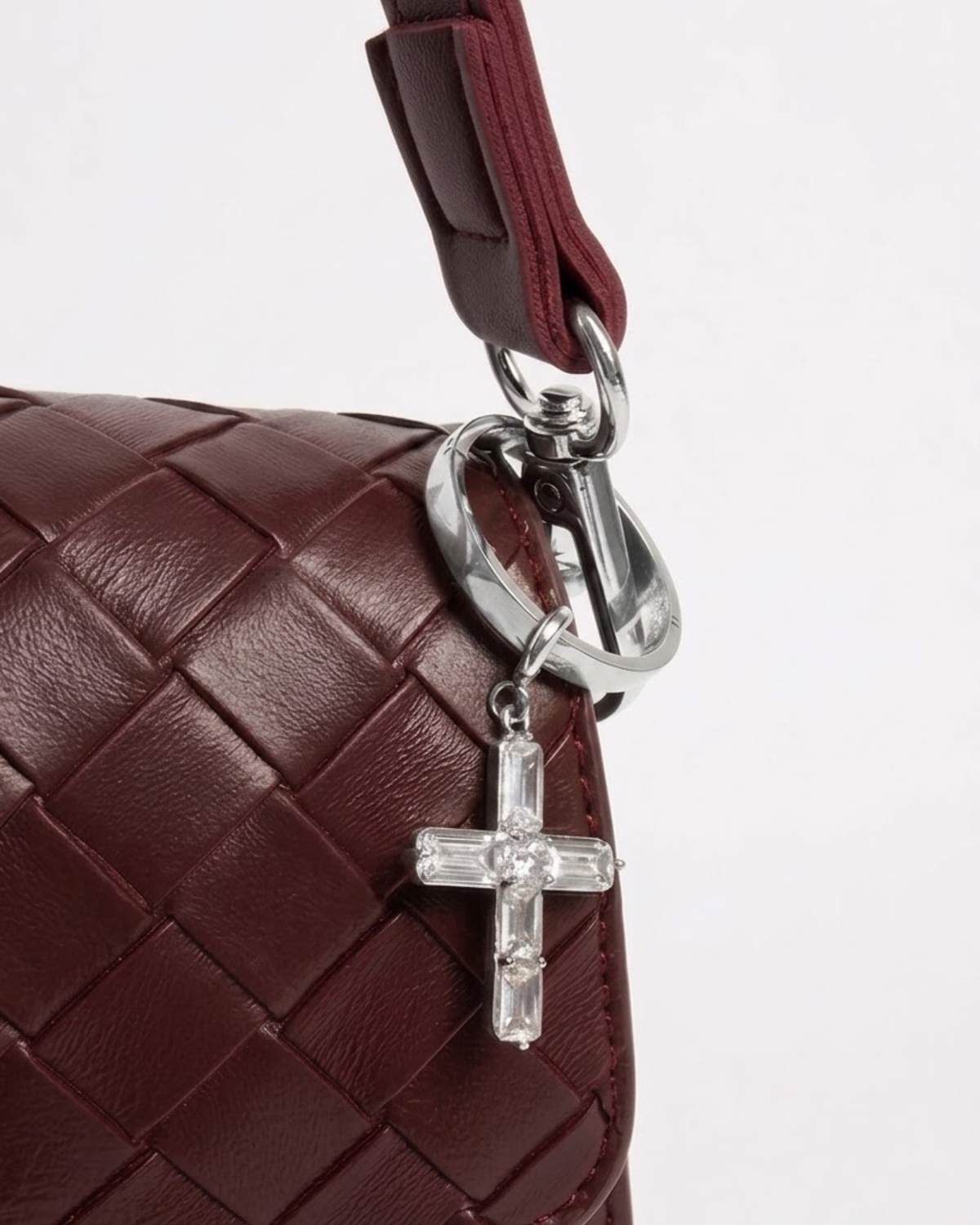 Silber BagBit -  Nahaufnahme einer weinroten, geflochtenen Ledertasche, an der ein Ring mit einem silbernen Anhänger in Form eines Kreuzes hängt, der mit klaren Schmucksteinen besetzt ist.