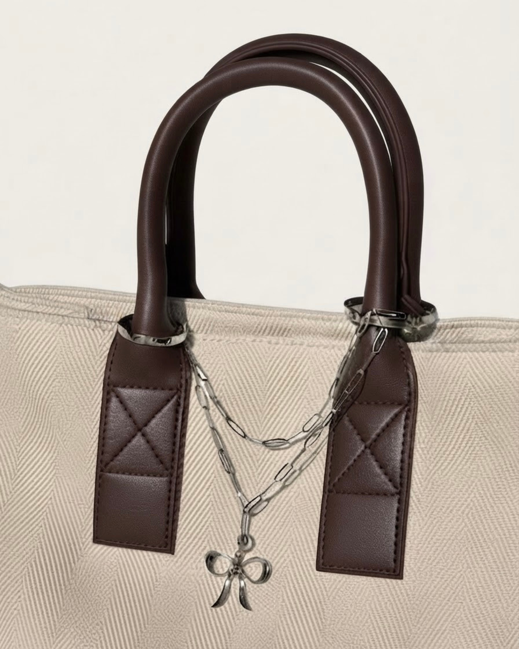 Silber Bag Chain - Beige-cremefarbene Tasche mit braunen Ledergriffen, verziert mit einer doppelreihigen, silberfarbenen Kette und einem zentralen Schleifenanhänger.