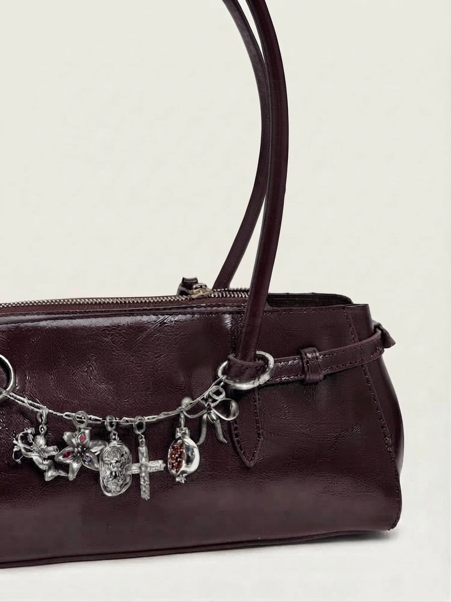 Silber Bag Chain - Dunkelrote Schultertasche aus glänzendem Leder, verziert mit einer silberfarbenen Kette zwischen den Henkeln und an dieser befinden sich mehrere silberne Anhängern (Motiv: Engel, Blumen, Kreuz, Schleife, Teddybär-Relief und Granatapfel).