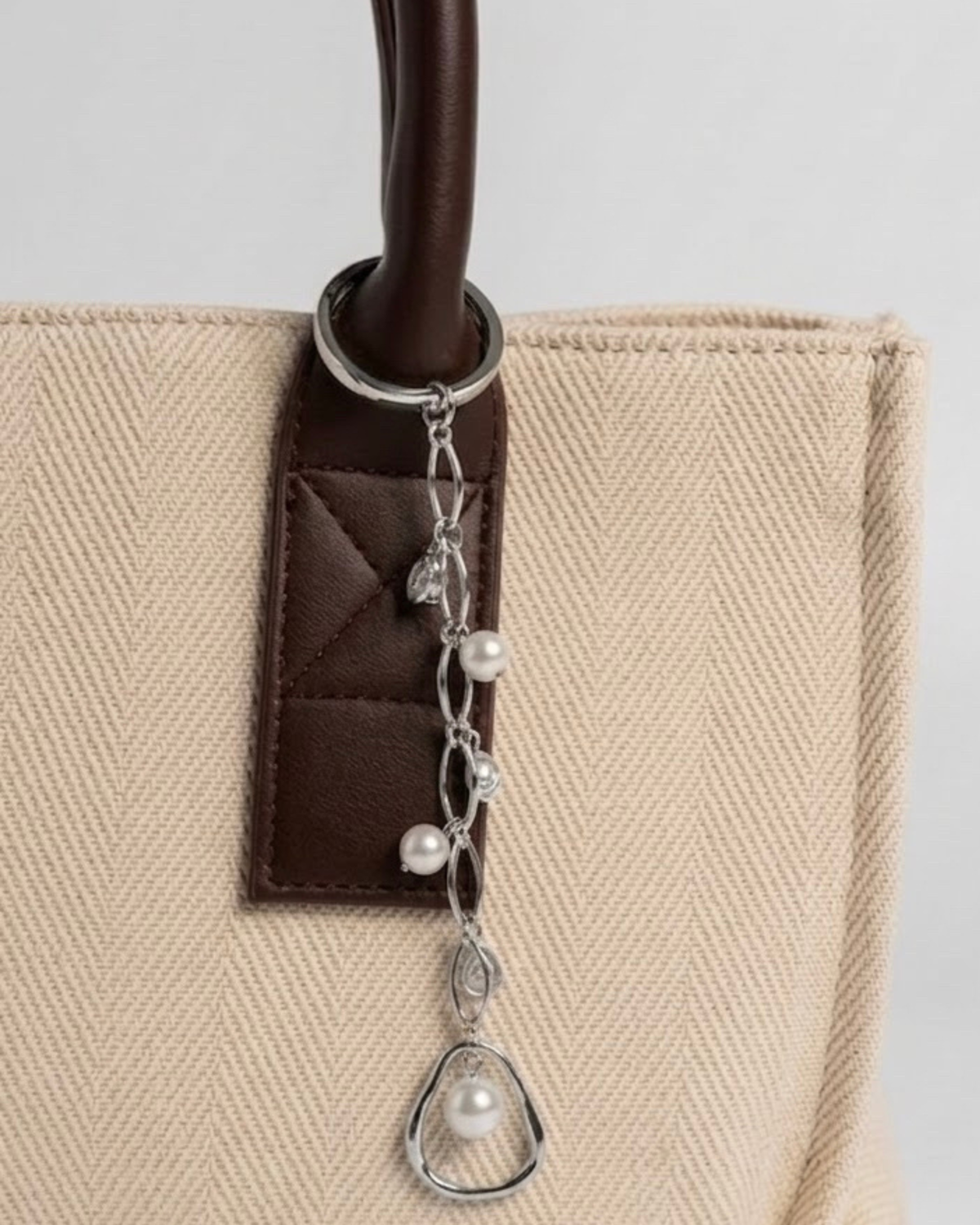 Silber Bag Charm -  Nahaufnahme einer beigefarbenen Canvas-Tragetasche mit braunem Ledergriff und einem filigranen silbernen Anhänger, der mit Perlen und tropfenförmigen Schmucksteinen verziert ist.