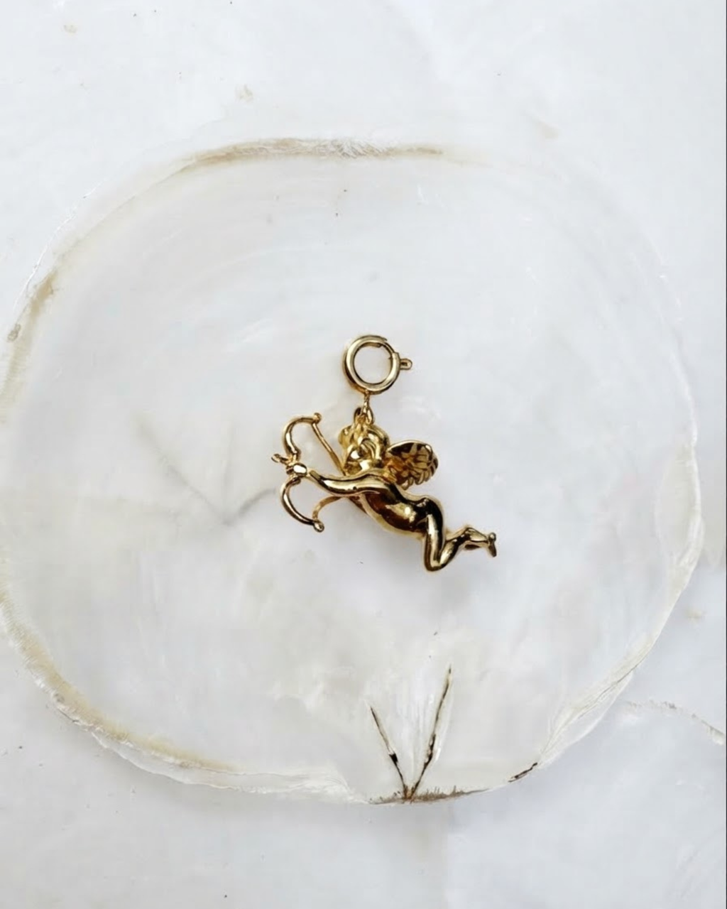 Gold Bag Bit - Goldfarbener Engel-Anhänger mit Pfeil und Bogen auf einer hellen Perlmutt-Unterlage.